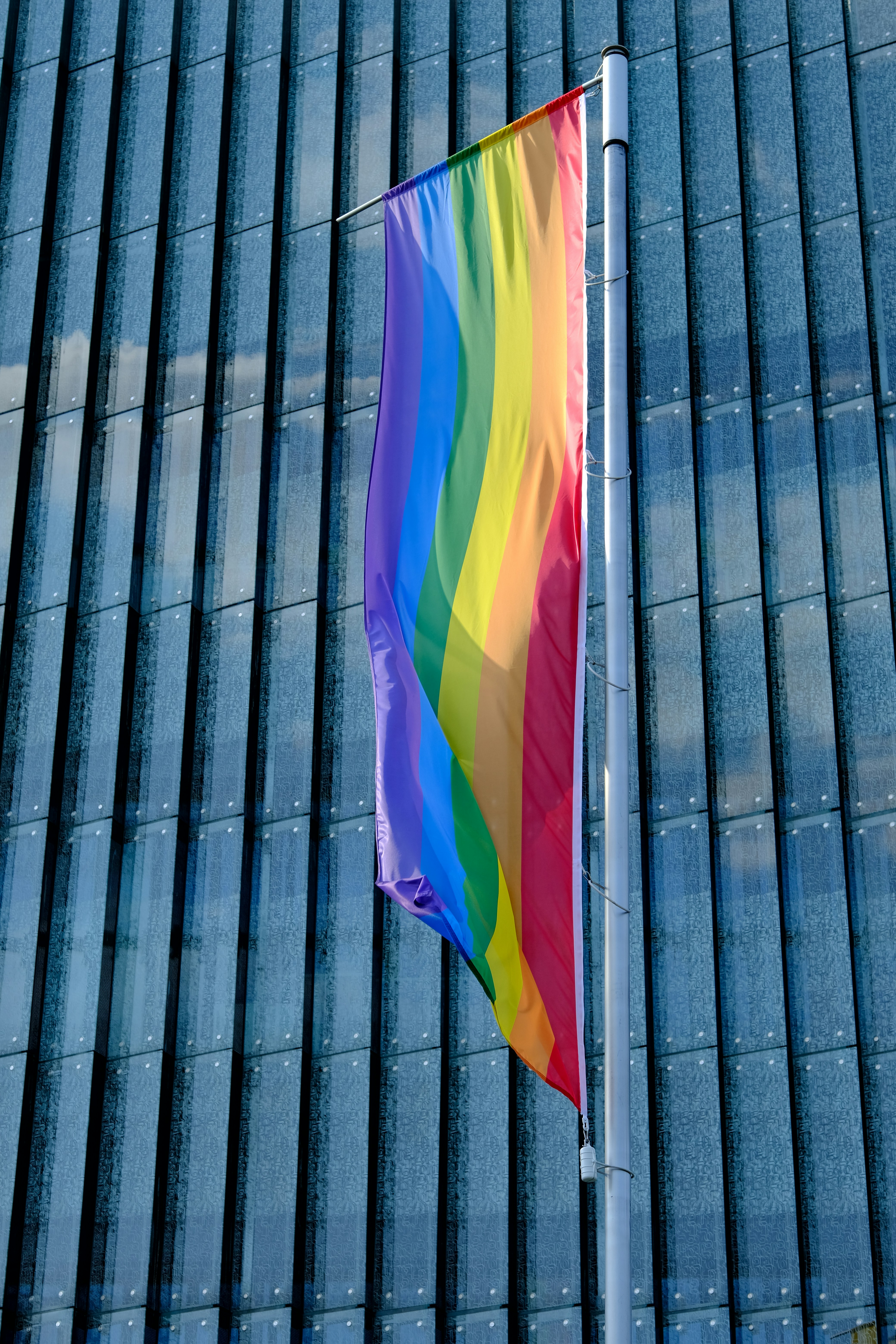 A pride flag hanging in the wind in front of a glossy skyscraper background.